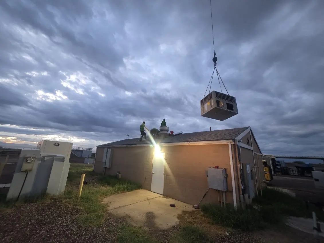 Rooftop air conditioning unit lifted by crane over a commercial building at dusk