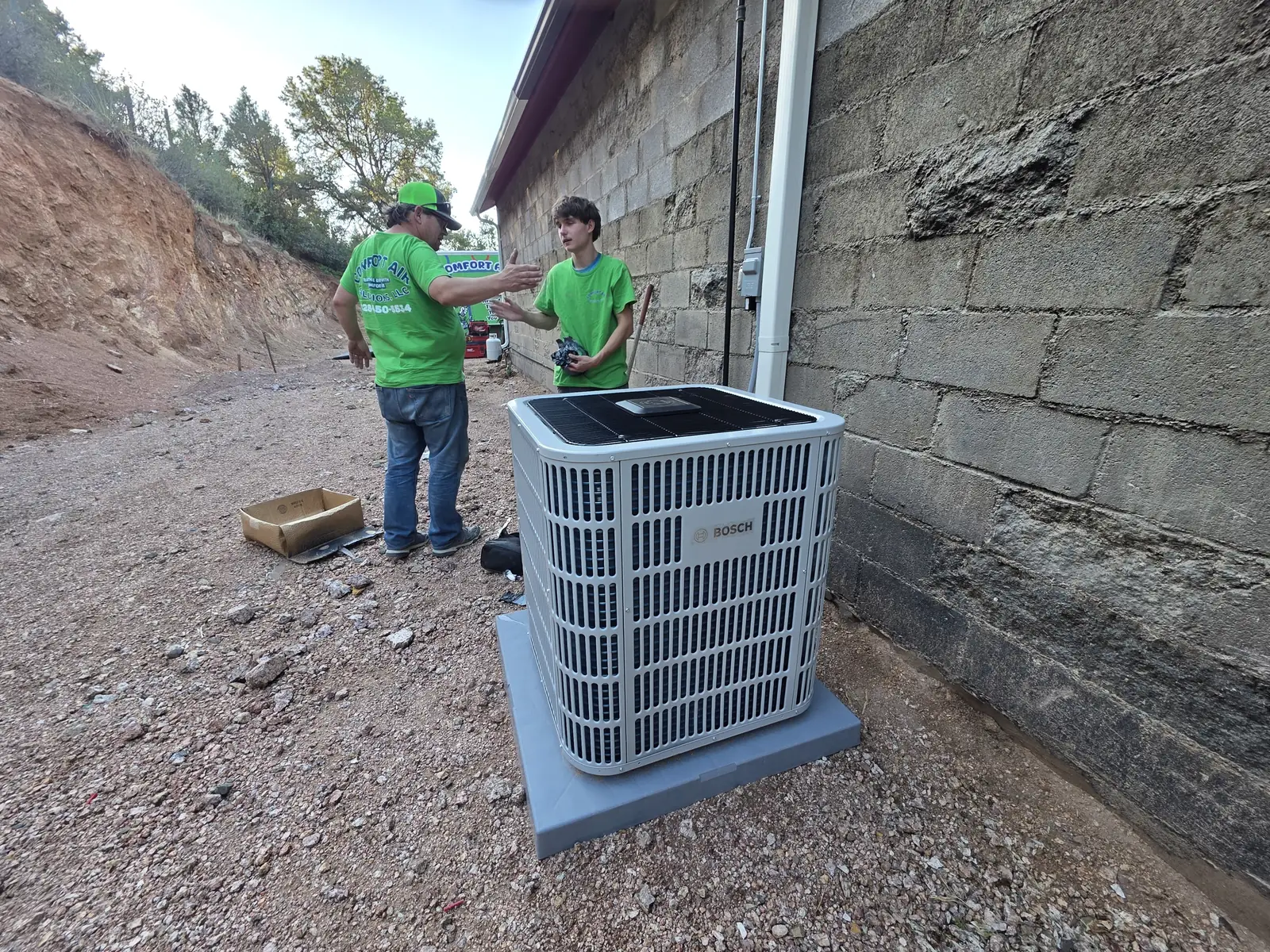 Comfort Air Solutions technicians setting a new outdoor condensing unit beside a block wall
